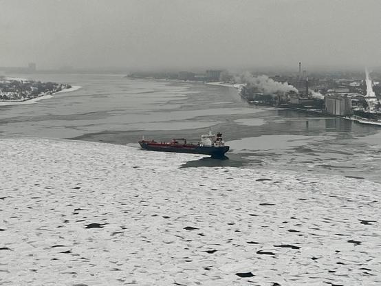 A cargo ship navigates through partially frozen water on the Detroit River, with ice patches visible. The background features a foggy Canadian skyline and smoke rising from industrial buildings. Photo credit to a family member.