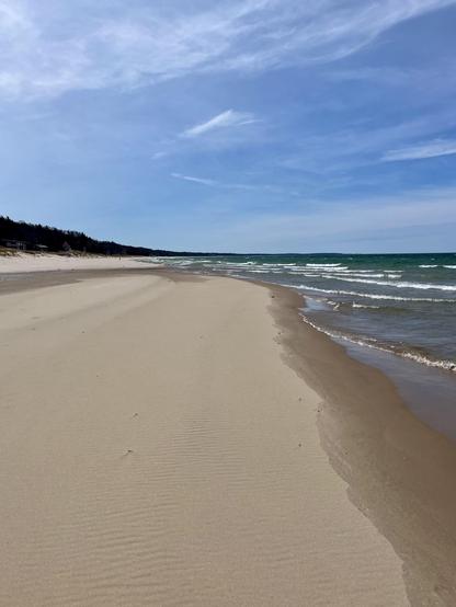 A sandy beach of Lake Michigan stretches along the shore, with gentle waves lapping at the water's edge. The sky is clear with soft clouds, and a tree line is visible in the distance on the dunes.