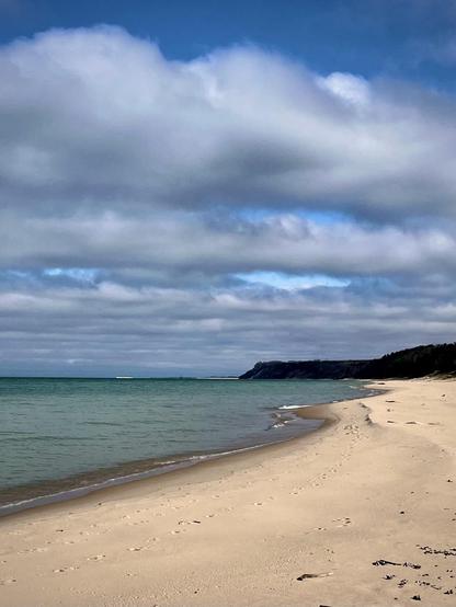 A serene beach scene with soft, sandy shores and gentle waves. The sky is partly cloudy, featuring varying shades of blue and gray. In the distance, a wooded shoreline meets Lake Michigan.