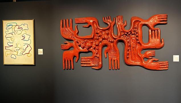 Two artworks on a wall - one seems to be a print or drawing with multicolored shapes; the other a 3-D red object with multiple hands emanating to various directions 
