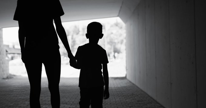 Silhouette of an adult holding the hand of a child while walking through a tunnel or archway, with light visible at the end. The image is in black and white.