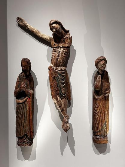 Three elongated wooden sculptures. On the left, the Virgin Mary, in the middle, Crucified Christ (one arm missing), and Saint John the Evangelist