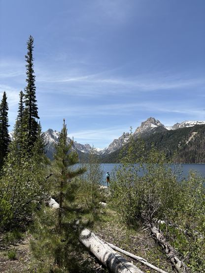 Sawtooth Mountains in Idaho landscape photo. Snow on the mountains, lake in between with trees in foreground. Streaky clouds in blue sky. My sweetheart  a small figure standing in the center with blue striped sweater. We love hiking!