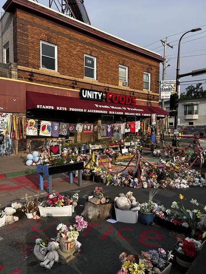 Store front, Unity Foods, and street with a variety of colorful memorials. One of the signs still says Cup Foods. 2025