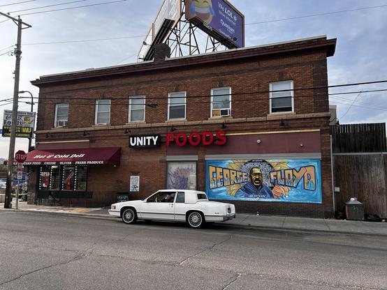 Side of a building with Unity Foods sign and iconic mural of George Floyd. A white car is parked on the street. 2025