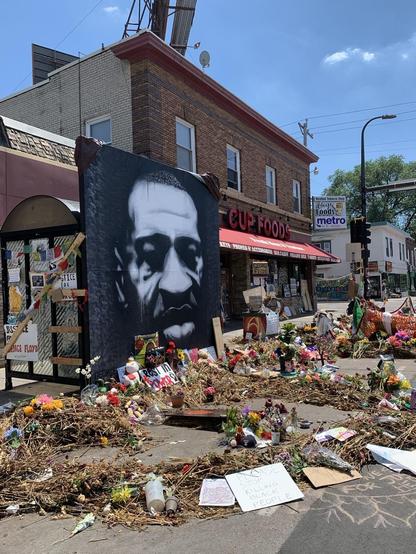 Storefront, Cup Foods, with large black and white image of George Floyd and colorful memorials on the street. 2020