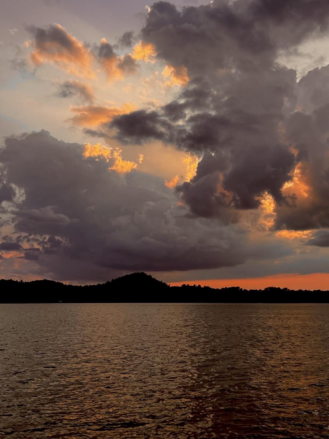 A serene sunset over a lake, featuring dramatic clouds with orange and peach hues illuminated against a darkening sky. Silhouetted hills outline the horizon.