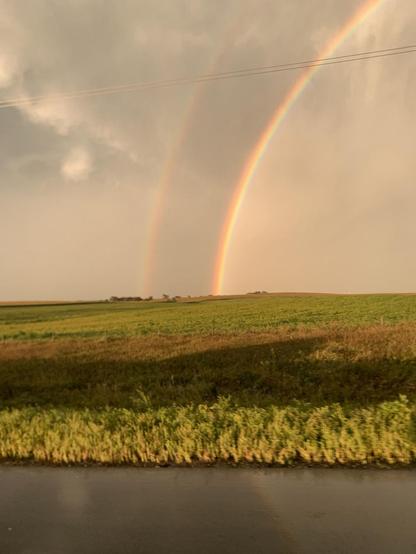 Cloudy sky with a double rainbow terminating in green farm fields with some water in the foreground. The image is fuzzy because it was taken from a moving car.
