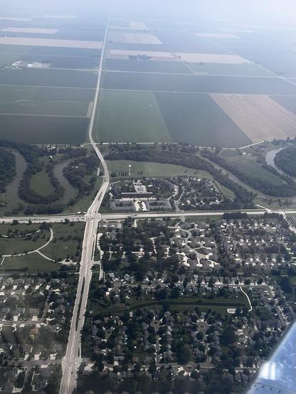 Aerial view of a suburban style town in the foreground, sinuous river, and green farm fields in the background. Two streets bisect the image.