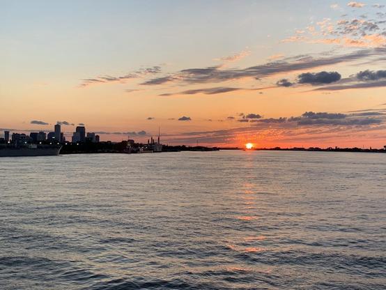 Expanse of water in the foreground of a setting sun, with silhouette buildings in the distance, a few clouds.