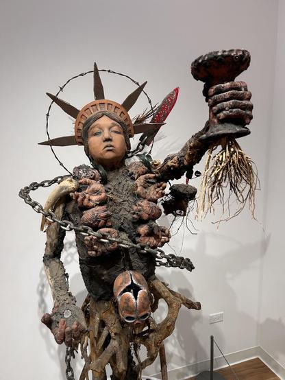 A mixed media sculpture that resembles the Statue of Liberty, but with barbed wire, chains, and body features 