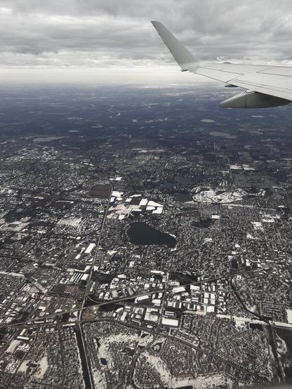 Suburban-ish development on low relief terrain with scattered small lakes. The snow cover gives roofs and grassy areas visual contrast. There's a portion of an airplane wing and low clouds. 