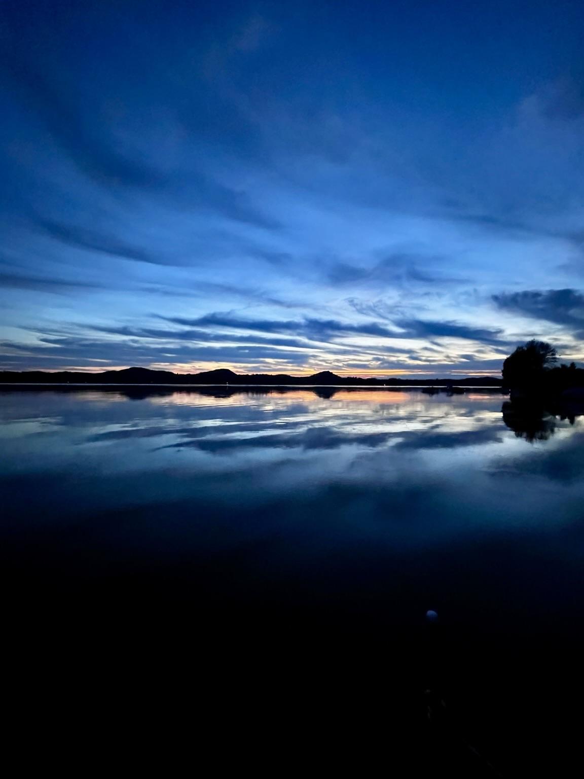 A wide angle view of a sunset over a lake as it transitions to night. The lake reflects the sky making the whole picture symmetrical from top to bottom. In the middle of the photo a thin row of black sand dunes are like beads on a string. Dark blue clouds create interesting patterns. A thin orange sunset betrays the last rays of the sun peaking over the horizon. I have been traveling this week so I pulled this photo from September.