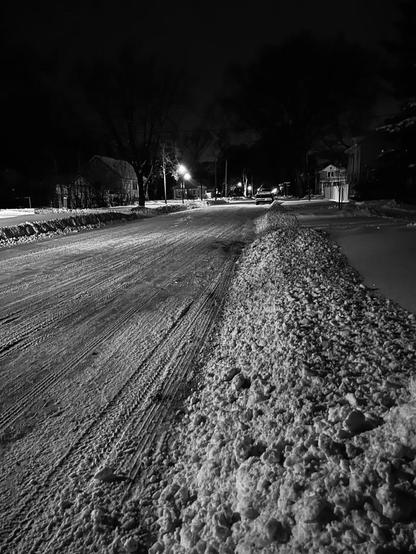 Black and white street view. A plowed, yet still snow covered, neighborhood street with snowbank on the boulevard. Cars, houses, and street lamps in the background.