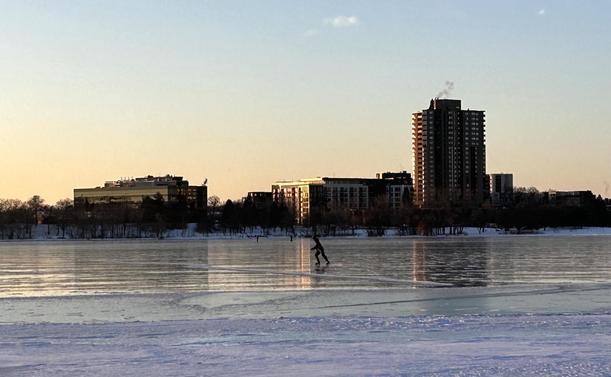 Frozen lake with an ice skater in the middle ground. Trees and buildings in the background under a clear sky.