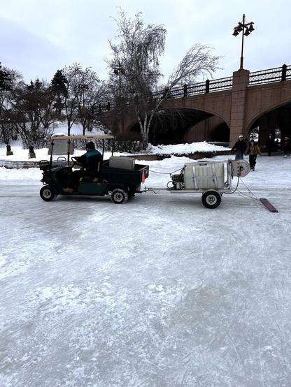 A person in a small, four wheeled vehicle, towing a trailer that is spraying water on an ice surface. Skaters and a bridge in the background.