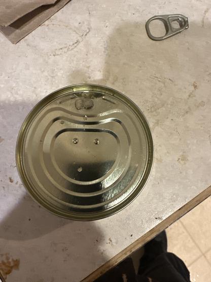 A small can (of Galician Tuna) with the pull-tab missing. The tab is sitting a few inches away.