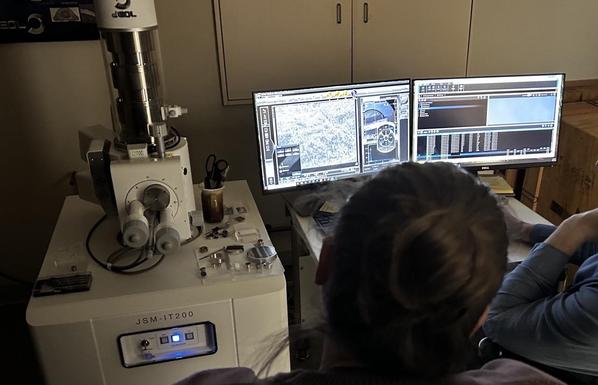 A JEOL JSM-IT200 electron microscope, with EDS X-ray detector. Next to it screens showing images of a sample surface and X-ray spectra. People are looking at the screens.