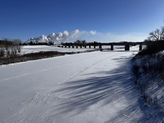 A flat expanse of snow, with snowmobile tracks, a bridge in the distance, a shallow bank on the left, and in the far distance a white plume drifting in a blue sky