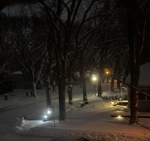 A dark, snow covered, tree lined, residential neighborhood. There are a few points of light, a couple of which are on a snowblower clearing a sidewalk.