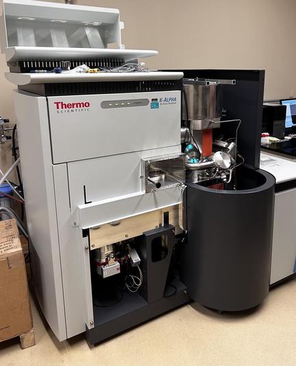 A floor standing instrument labeled Thermo Scientific and K-Alpha. Part of it is a sizable stainless steel vacuum chamber in which the sample is irradiated w X-rays, and the energies of ejected electrons are measured.