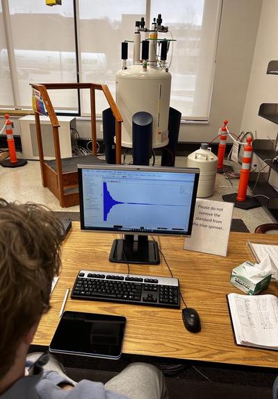 Lab setting. In the foreground, a person watching a computer screen, which shows a NMR (nuclear magnetic resonance) spectrum being collected. In the background, a Bruker NMR machine.