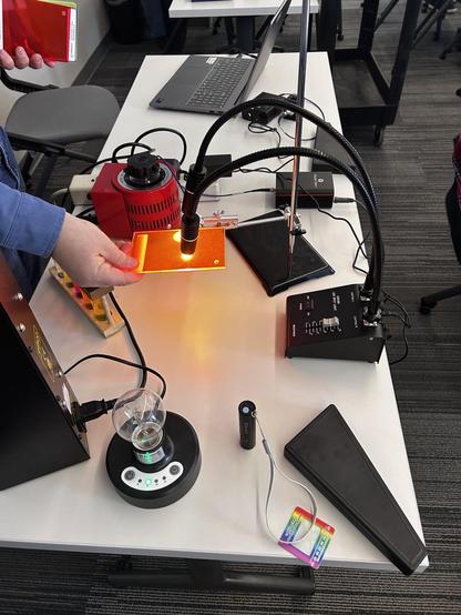 A table with apparatus, including a UV light source. A person is holding a piece of orange plexiglass under the source.