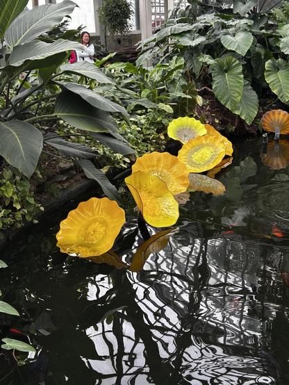 An indoor pool surrounded by lush plants, with large bright yellow glass flowers seemingly growing out of the water