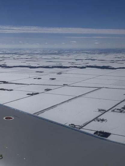 Foreground: an airplane wing; in the distance, flat, snow covered farmfields with scattered farmsteads, and roads cutting through at right angles. A sinuous tree lined river crosses the scene. The sky is blue with streaky high clouds.