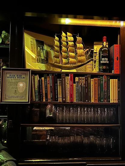 Shelves in the dark, nautical themed tavern, that include glasses, books, sailing ship decorations, and a note about the raw bar happy hour.