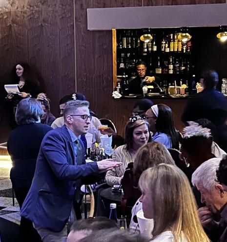 Inside scene of a club. A performer is doing sleight of hand for a seated group.