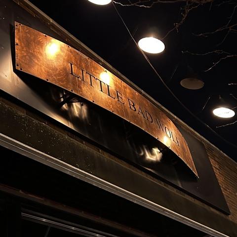 A curved brass colored sign over an entry that says LITTLE BAD WOLF. It's dark out and the sign is illuminated from above.