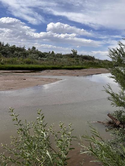 shallow river with a sandy opposite shore, trees and partially cloudy sky.