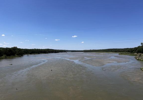 Looking downstream at a wide, shallow, river with a sandy bed. The shores are tree lined under a blue sky.