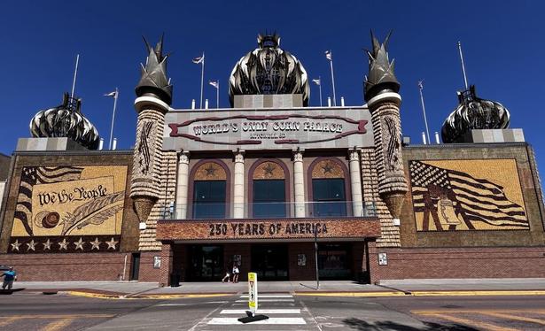 Façade and entry of a substantial building. On either side of the entrance are mosaics made of the halves of corn cobs. The entrance is flanked by turrets and onion domes. Signage: World's Only Corn Palace, Mitchell SD. 250 Years of America.