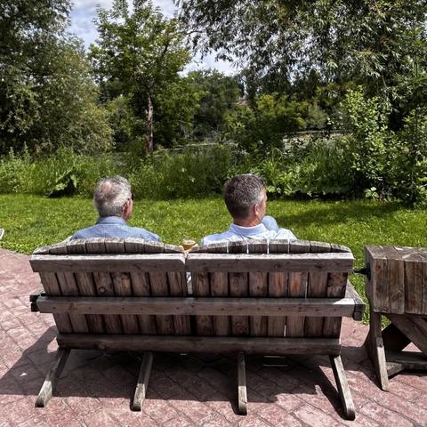 A couple of guys from the back, sitting in wooden chairs looking out over a quiet river.