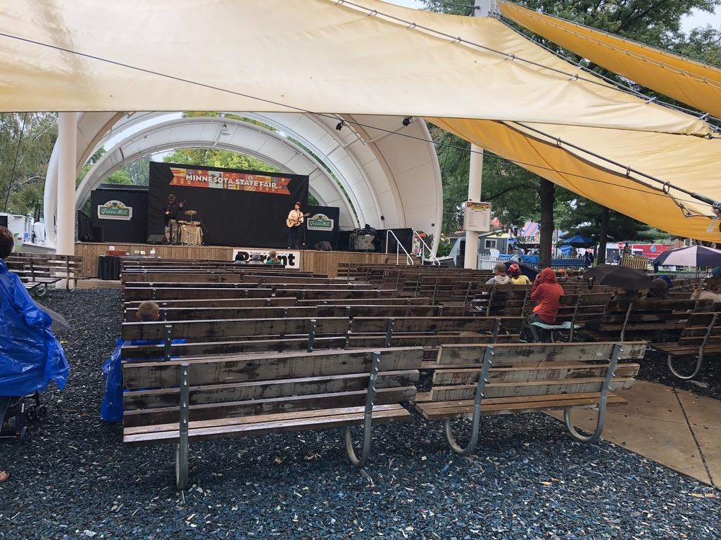 People in rain jackets sit under a tent at the Minnesota State Fair. About 10-12 people in attendence to see this folk duo play, sponsored by The Green Mill.