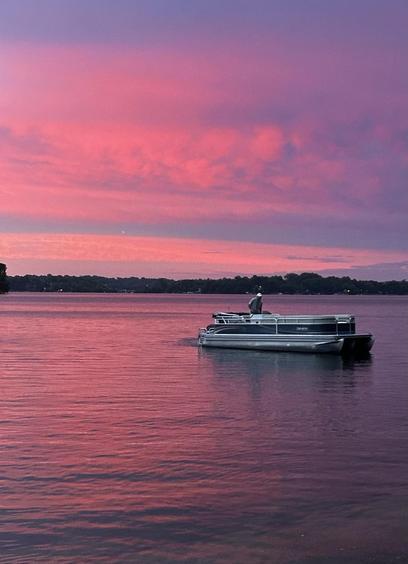Sunset over a lake, with pinkish colors reflected on the water. A person navigates a pontoon boat.