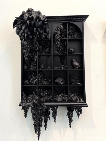 A black wall cabinet, filled and encrusted with shells and other items, all painted black. One of three.