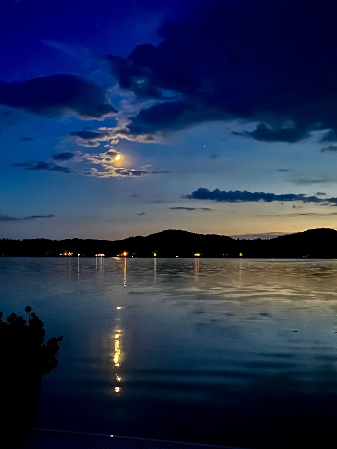 52 minutes after sunset. It’s all about the blues. And the moon. View over a calm lake. Along the horizon a gentle yellow sunset is still visible, but the colors quickly transition to teal, dark blue, and purplish blue. All the blues. The colors are reflected on the barely undulating lake surface which almost looks like ice but it is still hot outside. There are dark clouds on the upper right. The star of the show is a bright moon partially hidden behind small clouds which are illuminated by the moon in a mysterious way. The moonlight is also reflected in a pattern of dots on the lake surface towards your eye. In this way the hidden sun still finds its way to you by bouncing off our moon and the water. On the distant shore small lights are visible through the trees with silhouetted sand dunes as the back drop. 